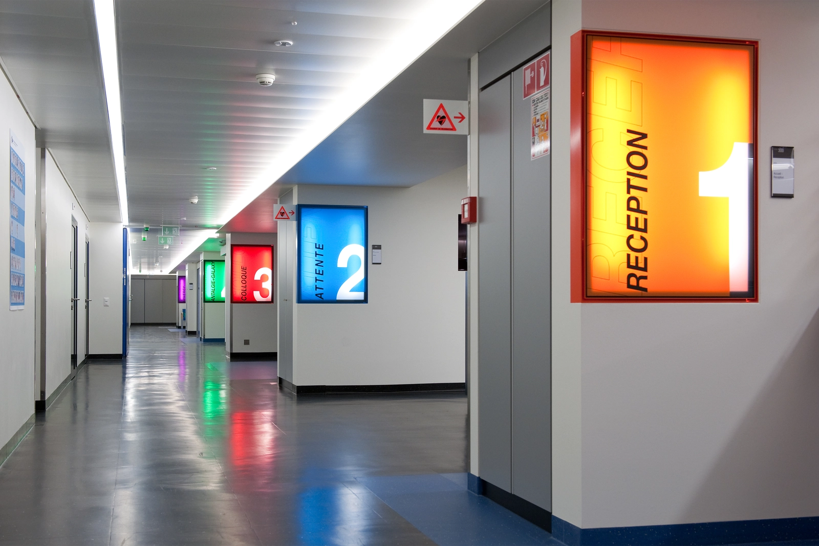 Photo d'un couloir avec des panneaux colorés qui indiquent les différents lieux "réception", "attente"